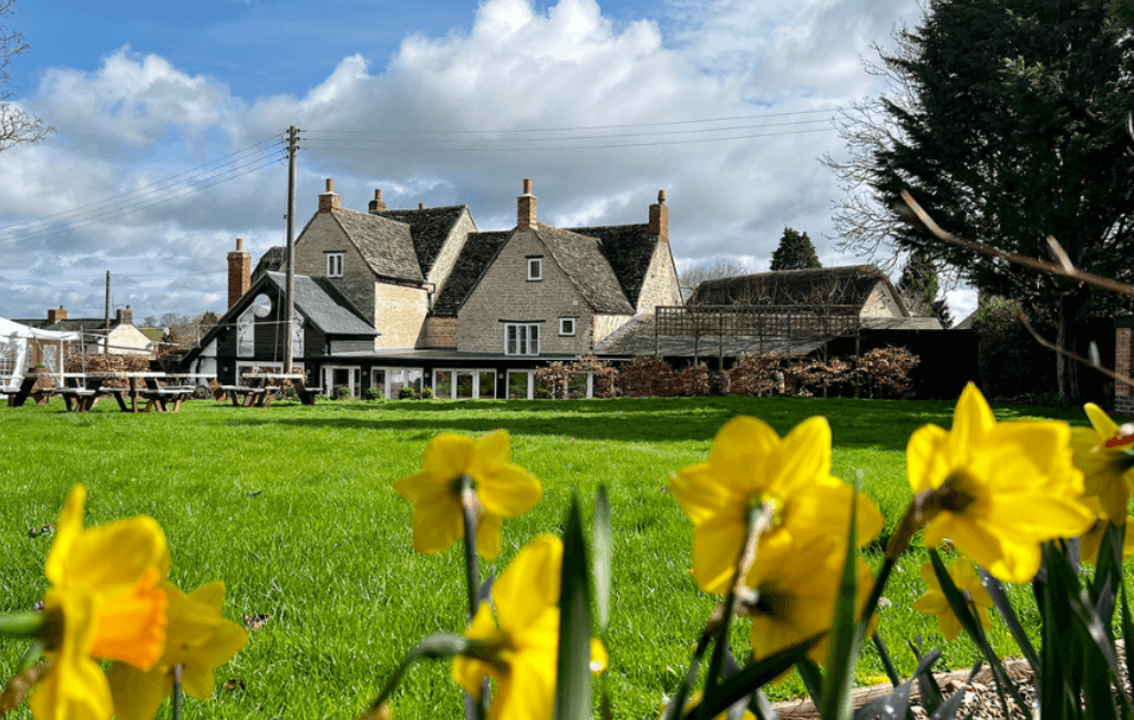A cottage is shown in the landscape and in front of the image there are some yellow flowers