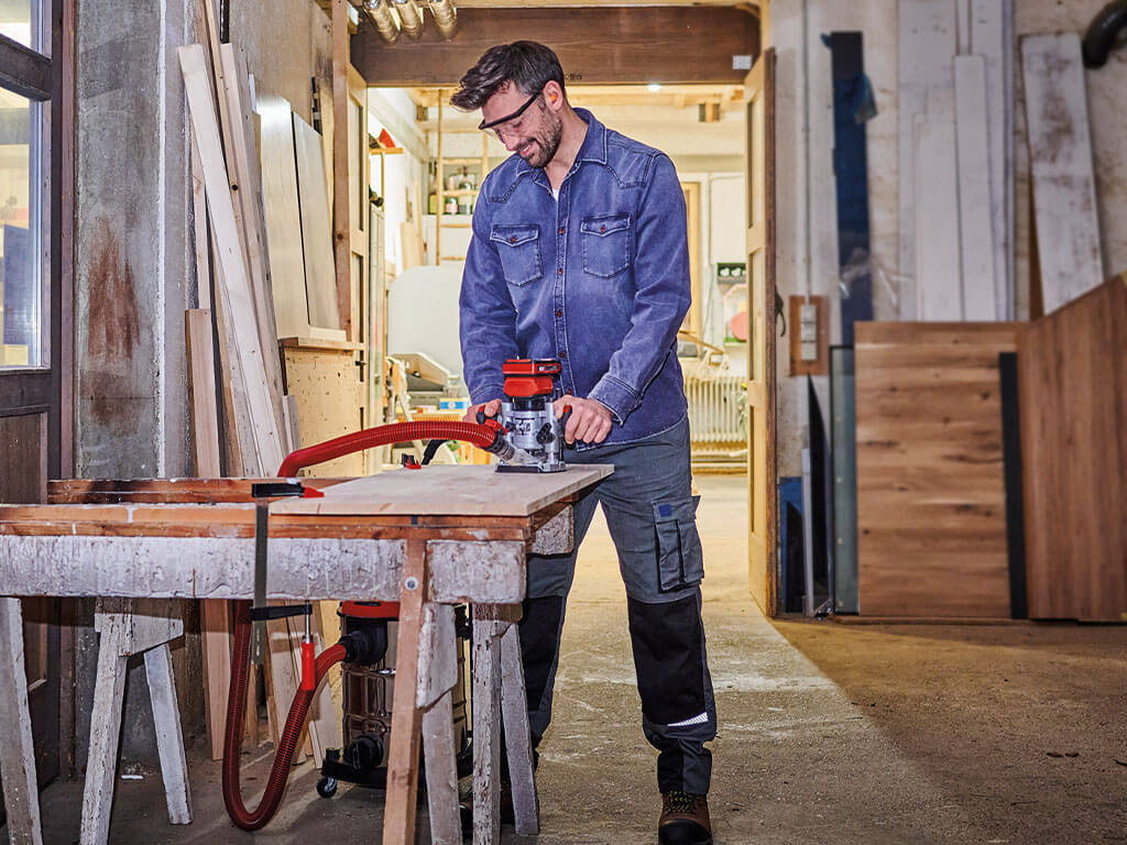 A man is standing in his workshop working on a wooden panel with the Einhell cordless router and a suction device.