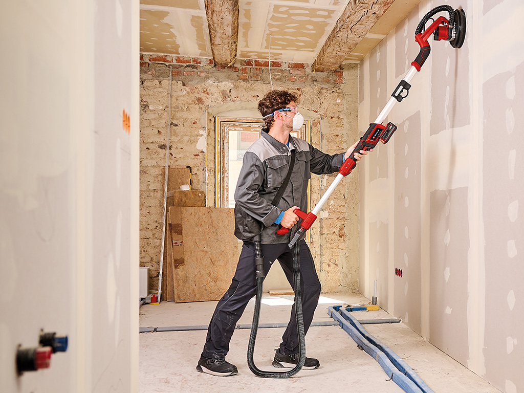 A man sands the wall with a drywall sander.