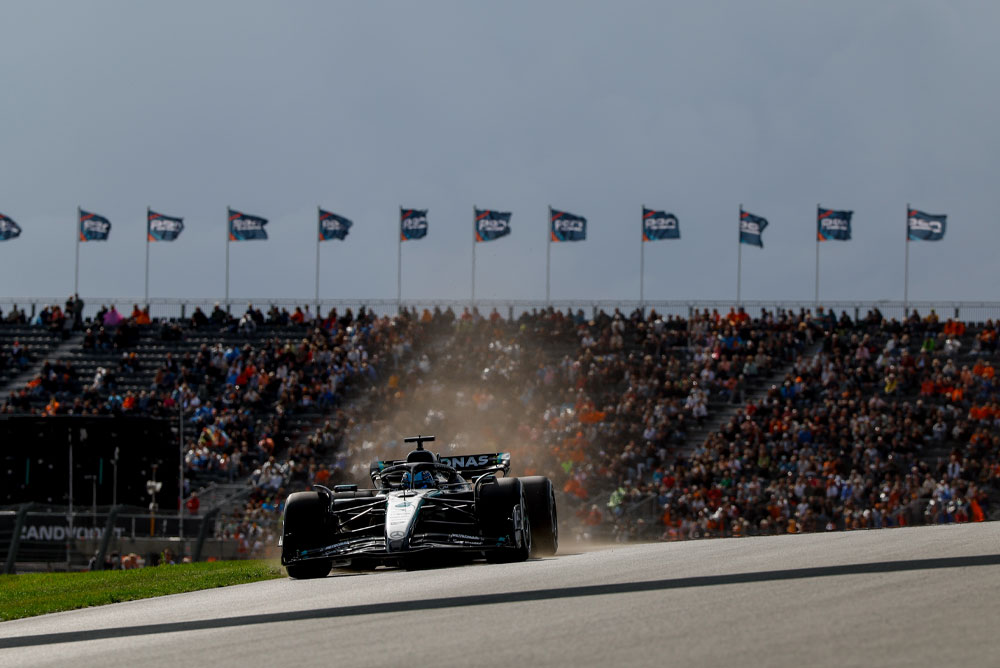 A Mercedes-AMG PETRONAS Formula One car drives past packed grandstands during a race. Fans wave flags in the background, creating a vibrant atmosphere.