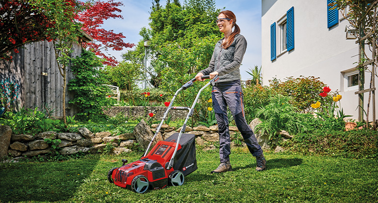 A woman using a einhell scarifier with a catch bag to work on the lawn in a well-kept garden with flower beds and a white house in the background.