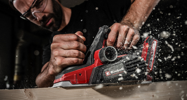 A cordless planer smooths a wooden surface, producing fine wood shavings.
